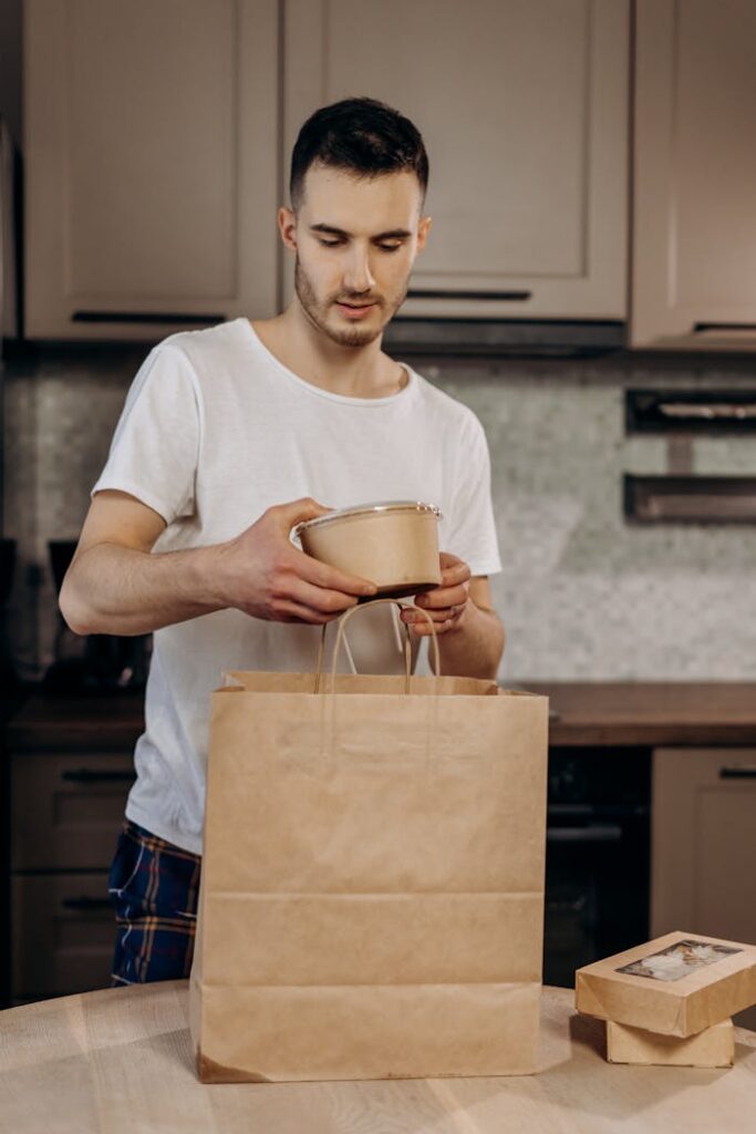 A young man in a kitchen packing takeout food into a brown paper bag, showcasing eco-friendly packaging.